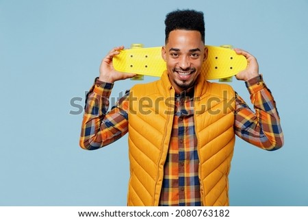 Smiling businesslike charismatic fun young black man 20s years old wears yellow waistcoat shirt hold skateboard behind head on shoulders isolated on plain pastel light blue background studio portrait