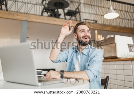 Joyful young man sit at table in coffee shop cafe restaurant indoors working studying on laptop pc computer waving greeting with hand as notices someone. Freelance mobile office business concept Royalty-Free Stock Photo #2080763221