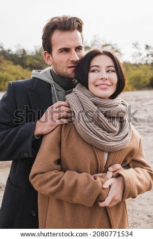 young man hugging cheerful woman in coat and scarf during autumn walk