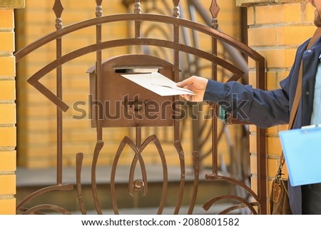 Handsome young postman putting letter in mail box outdoors Royalty-Free Stock Photo #2080801582