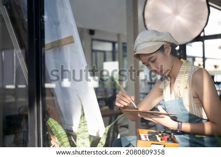 Pretty young woman holding palette and paintbrush while working on painting in her workshop.