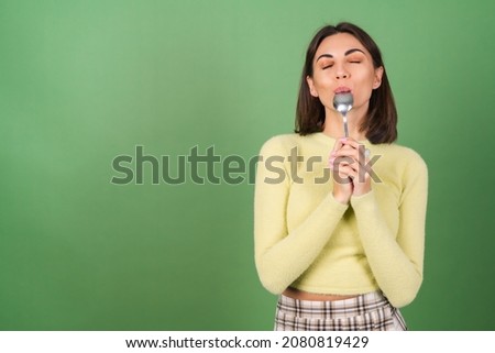 Young woman on a green background in a yellow sweater with appetite eats from a tablespoon, enjoying Royalty-Free Stock Photo #2080819429
