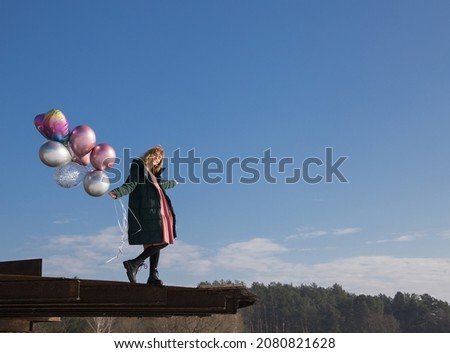 full-length portrait of gentle graceful teenage girl holding bunch of colorful balloons in hands. Stands high on roof against blue sky on sunny day. feels great, festive, magical, dreamily joyful