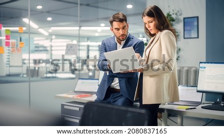 Young Manager Talking with Experienced Colleague while Using Laptop Computer in Office. Colleagues Discuss Business, Financial and Marketing Projects. Specialists Work in Diverse Team. Royalty-Free Stock Photo #2080837165