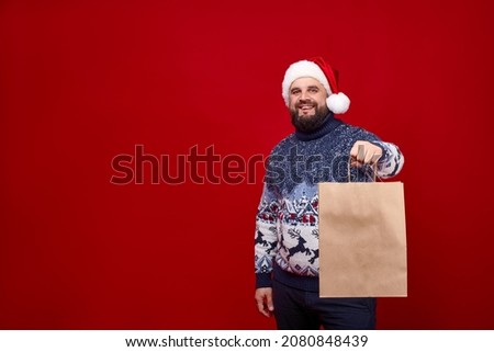 A bearded man in a Santa hat and a sweater with deer shows a craft package to the camera on a red background. A bearded guy in a sweater with deer looks at the camera. Space for logo on shipping bag