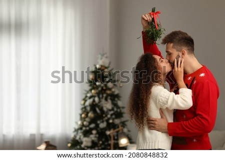 Happy couple kissing under mistletoe bunch in room decorated for Christmas