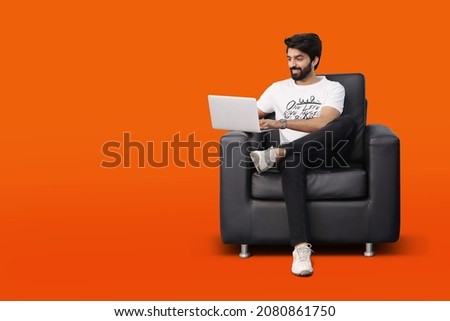 Happy young bearded man sitting on black sofa couch chair using laptop on orange background - Pakistani Indian South Asian