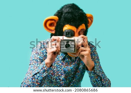closeup of a young man, wearing a monkey mask, taking a picture with a retro instant camera on a blue background