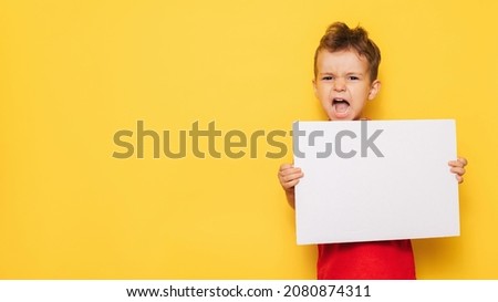 Studio portrait of a screaming boy with a clean white poster in his hands on a bright yellow background, with a place for your text or advertising.