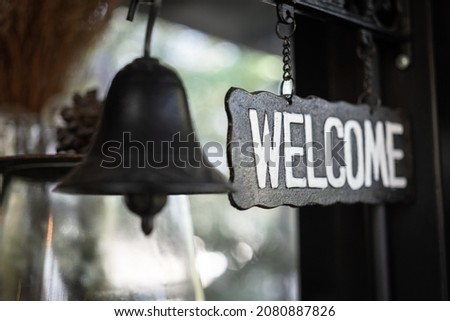 "Welcome" in white text color on rustic style metal sign plate which is hanged with bell. Interior decoration object photo. Close-up and selective focus.