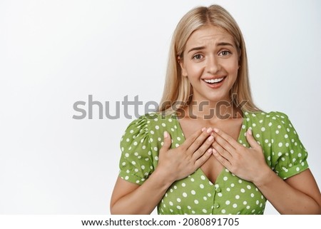 Cute emotional girl looking flattered at camera, holding hands on chest and smiling, express gratitude, standing in dress against white background