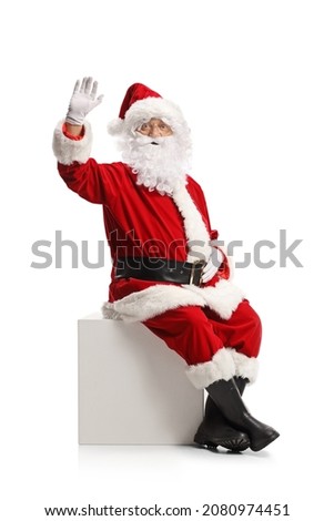Santa claus sitting on a white cube and waving at camera isolated on white background