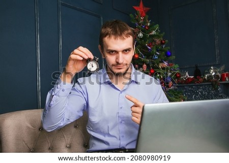 A man works at a laptop on New Year's Eve against the background of a Christmas tree. Employment in Christmas conicles. holding a watch