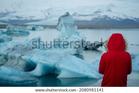 Rear view of female tourist, young woman in red jacket with a hood  looking at floating ice in Jokulsarlon glacier lagoon in Iceland, enjoying amazing view on a sunny day