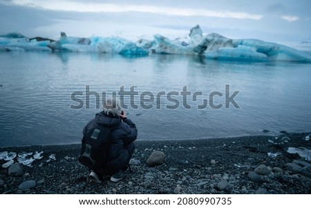 Old Photographer takes a picture in Jokulsarlon, Iceland with a small iceberg floating in the lake. Epic travel in a wilderness