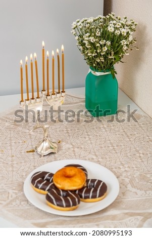 Hanukkah candlestick with all burning candles and donuts and a vase of flowers on the table. Vertical photo