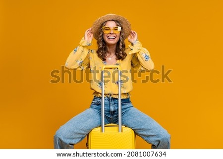 stylish happy smiling blond woman traveling with suitcase dressed in trendy outfit jeans, blouse and hat, sunglasses, bright yellow background, positive summer vacation, fashion trend Royalty-Free Stock Photo #2081007634