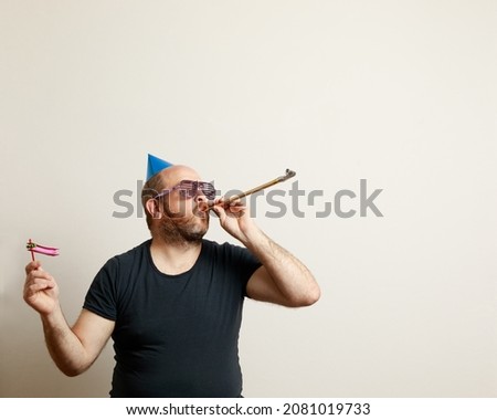 A Portrait of Caucasian man celebrating joyfully. He has birthday hat, rattle, bugle and funny cotillion glasses. Neutral background. Horizontal image with copy space for text.