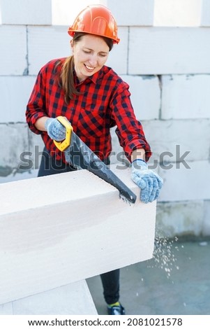 Portrait of a young attractive female construction worker in a protective helmet, sawing aerated concrete blocks with saw. process working bricklayer's helper, preparing block for laying. Copy space