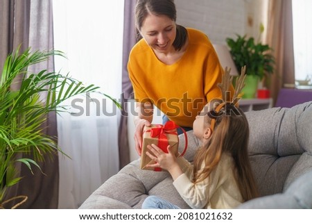 Loving mom gives her daughter gift for Christmas. little girl with deer horns on her head is happy and laughing. hugging and holding Christmas gift box against background garland lights.