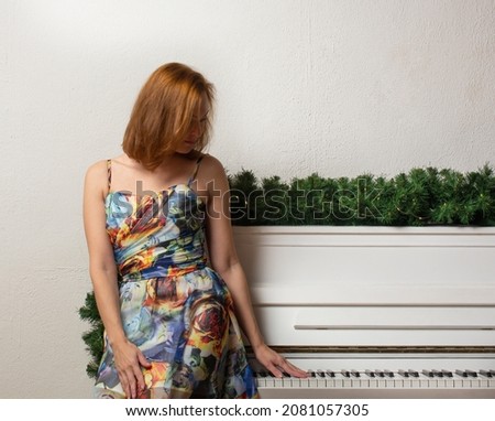 a girl with red hair in a blue and white dress near the piano and a Christmas tree against a white concrete wall. copy space.