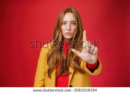 Serious-looking redhead woman in yellow coat extending hand to camera with stop and warning gesture prohibiting and forbidding make something having unemotional face as being confident over red wall