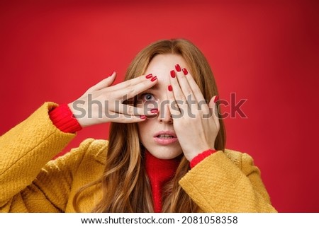 Stunned and shocked redhead woman cannot believe what terrible thing she sees covering face with palms open mouth from shook and peeking through fingers anxious and scared over red background