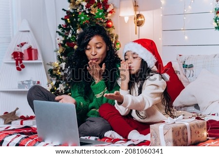 African American woman and brunette daughter in Santa hat sit among Christmas decorations and blow kisses chatting on video call via laptop