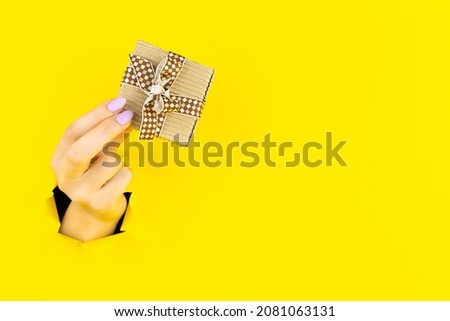 Female hand giving present, gift, surprise in torn yellow paper hole background. Breaking on, breakthrough. Concept of celebration, shopping, proposal, sales, ad. Copyspace. Promotions discounts.