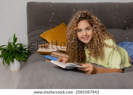 blonde girl with blue eyes and curly hair is reading a book lying on a gray bed. concept of education. home reading. space for text. High quality photo