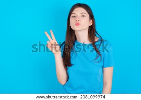 Young caucasian girl wearing blue T-shirt isolated over blue background makes peace gesture keeps lips folded shows v sign. Body language concept