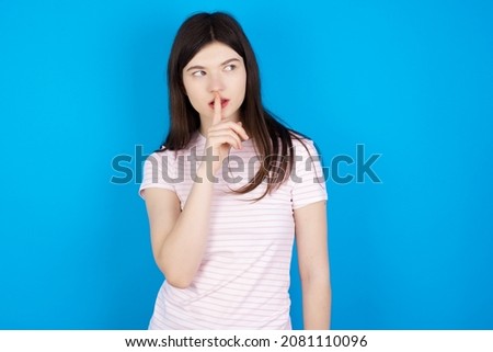 Caucasian woman wearing striped T-shirt over blue background silence gesture keeps index finger to lips makes hush sign. Asks not to share secret