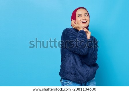 Portrait of smiling young asian woman with hand on cheek and looking to side on blue background