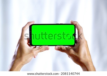A man holds a green screen smartphone with both hands in Landscape Mode and white background. product placement.