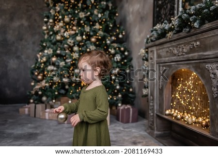 Close up portrait of little girl in warm green dress holds golden ball in hands, stands by bright wooden fireplace near the Christmas tree with gifts. Decorating a stylish holiday apartment interior