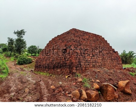 Stock photo of huge pile of brown or red color bricks in the construction site surrounded by green trees at rural kolhapur, Maharashtra, India. Picture captured during monsoon season. selective focus.