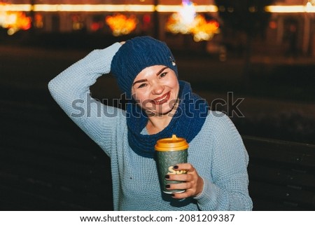 Photo of attractive cheerful woman sitting on a bench in cold snowy weather, smiling, drinking coffee outdoors
