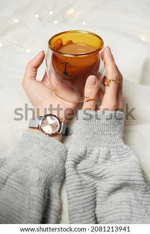 Shot of female hands holding a cup of coffee. The girl wears a pullover, watch, rings. The inside of the double-bottom cup is yellow and shaped as a teddy bear. The items are on the white background.