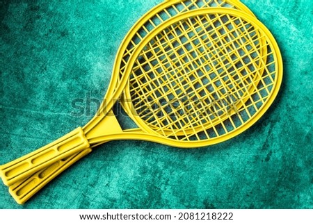 two plastic tennis net rackets of bright yellow color are laid out diagonally on a worn turquoise background; the children's version of the tournament is badminton