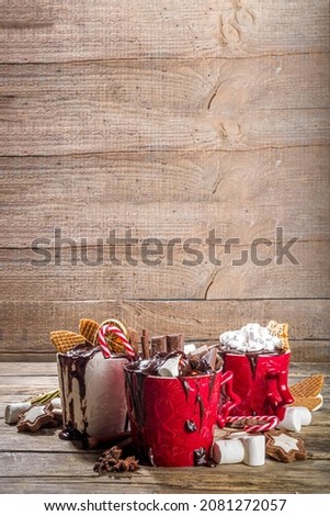 Christmas styled overloaded hot chocolate mugs with various toppings – chocolate slices, spices, sweets, cookies, candy and gingerbreads, wooden cozy winter background copy space