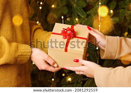 Female hands holding craft gift box with red ribbon near Christmas tree decorated with lights. New year presents