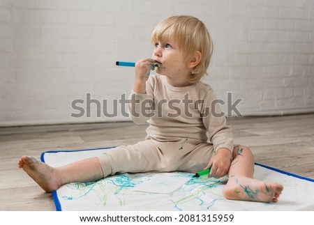 A little cheerful girl in pajamas is sitting on the floor and dreamily gnawing on a water marker. The face and clothes are stained with paint
