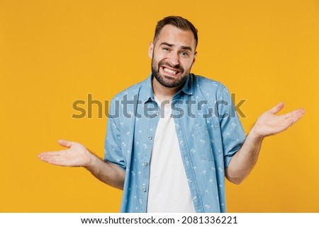 Young confused sad man 20s wearing blue shirt shrugging shoulders looking puzzled, have no idea, nothing to say, standing questioned and unaware isolated on plain yellow background studio portrait.