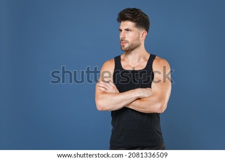 Attractive young bearded fitness sporty guy sportsman in black undershirt isolated on blue background studio portrait. Workout sport motivation lifestyle concept. Holding hands crossed, looking aside