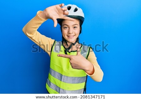 Beautiful brunette little girl wearing bike helmet and reflective vest smiling making frame with hands and fingers with happy face. creativity and photography concept. 