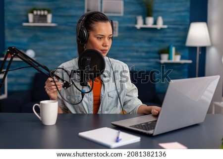 Woman vlogger streaming podcast and using laptop at desk. Influencer with headphones in home office recording livestream on computer. Blogger live broadcasting for social media with microphone. Royalty-Free Stock Photo #2081382136