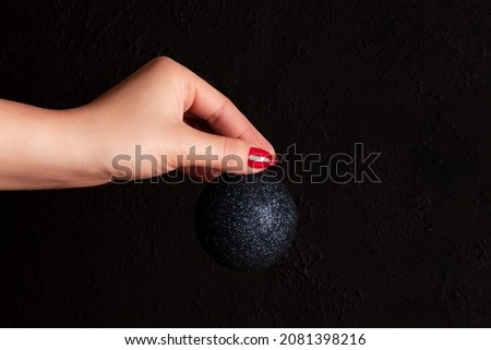 Female hand with blue glittery Christmas ball with beautiful manicure - red nails against black background. Nail and skin care concept
