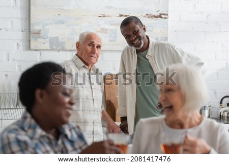 Senior multiethnic men looking at camera near women holding tea in nursing home