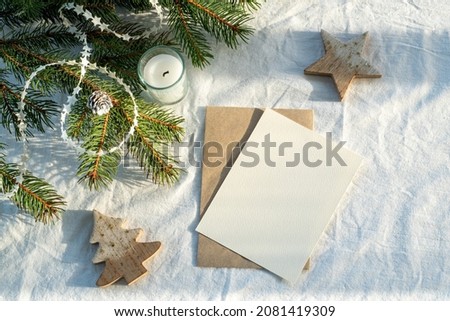 Blank greeting card, invitation card mockup.Christmas decoration scene in sunlight with card, envelope, ribbon, fir branches, candle and wooden balls.White linen background.Top view, flat lay.