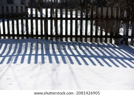 A beautiful contrasting blue shadow of a rustic village fence on the white snow on a sunny winter day. Background. Abstraction.